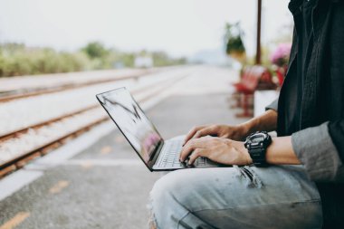 Genç Asyalı adam dizüstü bilgisayarını tren istasyonunda çalışmak ve seyahat yeri aramak için kullanıyor. Serbest çalışma ve seyahat kavramı.