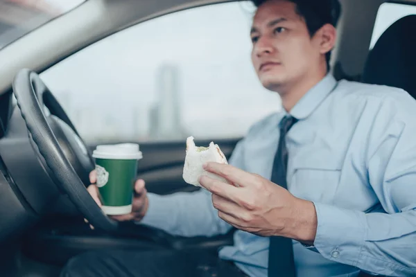 The businessman is eating a sandwich and hold cup of coffee while driving on the road, and he is not without a seat belt.