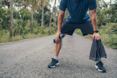 Yorgun genç bir adam dinleniyor ve nefes alıyor. Elinde bir şişe içme suyu ve havlusu var. Sabah parkta yolda..