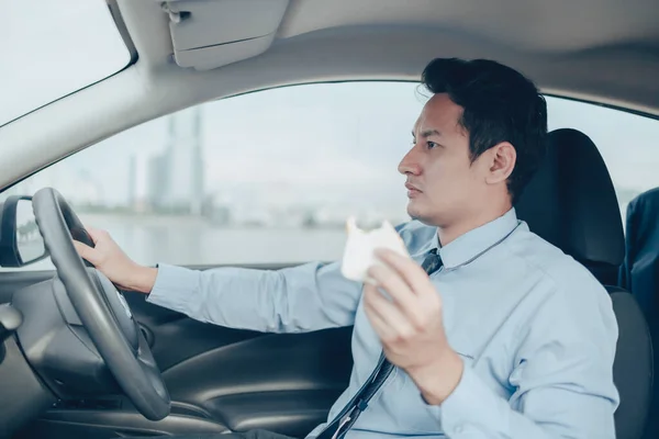 The businessman is eating a sandwich while driving on the road, and he is not without a seat belt.