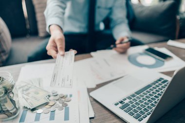 Ofisteki masada müşteriye ve dolar, para, dizüstü bilgisayar ve grafik grafiğine çek veren bir iş adamı. Çek ile ödeme, maaş çeki, maaş kavramı.