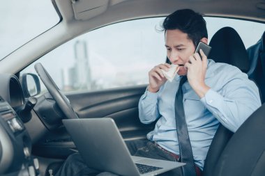 İş adamı laptopta çalışırken sandviç yiyor ve arabasında sürücü koltuğunda telefonla konuşuyor. Meşgul işadamı ve arabada yemek