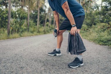 Yorgun genç bir adam dinleniyor ve nefes alıyor. Elinde bir şişe içme suyu ve havlusu var. Sabah parkta yolda..