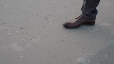 Ayakkabılı ve pantolonlu bir adamın ayakları sahilde yürüyor.