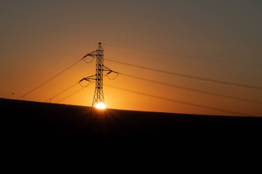 Güneş bir elektrik kulesinin ortasında saklanıyor ve arka planda ışığa karşı bazı dağlar var. Kule görüntünün solunda..