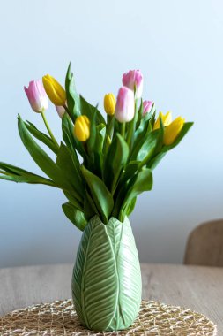 Vertical photo of a beautiful composition of pink and yellow tulips inside a green vase on a wooden table. White background and selective focus.