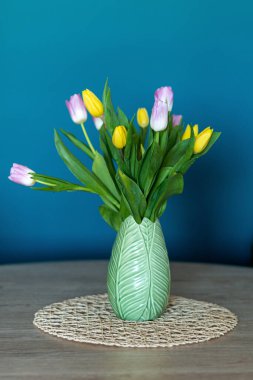 Vertical photo of a nice composition of pink and yellow tulips inside a green vase inside my house. Oil blue background.