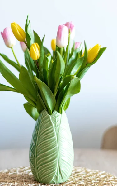 Vertical photo of pink and yellow tulips inside a green vase inside my house. White background and selective focus.