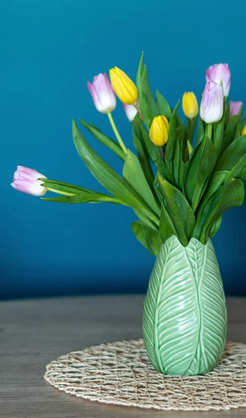 Vertical photo of a nice composition of pink and yellow tulips inside a green vase inside my house. Oil blue background and selective focus.