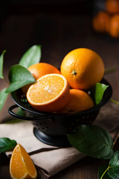 A few juicy oranges in a black bowl with green leaves on a dark wooden background. Dark, rustic and close up concept.