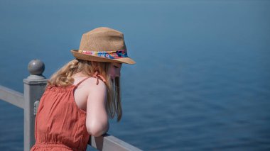 Bir kız deniz kenarında durur ve güneşli bir yaz gününde yüzsüz bir şekilde rüya gibi uzaklara bakar.