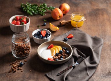 Sağlıklı ve besleyici kahvaltı kavramı. Ev yapımı tahıllı lezzetli yoğurt; şeftali; çilek; bal; ve koyu ahşap arka planda bir kasede yabanmersini. Diyet yemek..