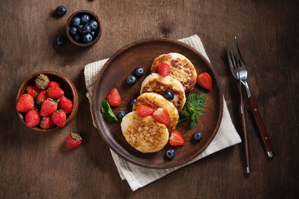 Cottage cheese pancakes or syrniki with blueberries, strawberries and fresh mint on a dark wooden background. The concept of a home-made healthy breakfast. Top view image.