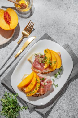 Şeftalili sandviç, krem peynirli, jambonlu ve ballı kekik, meyve ve gölgeli gri bir tabakta. Reklam menüsü ya da kafe afişi için sağlıklı beslenme atıştırmalığı konsepti. Üst görünüm, alanı kopyala.