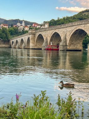 Huzurlu Drina Nehri 'ni çevreleyen kemerli, ön planda yabani çiçekler ve arka planda yeşil tepeler ve evler olan antik bir taş köprü. Miras ve gelişen turizm konsepti. Visegrad, Bosna-Hersek.