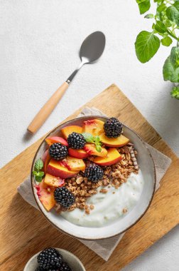 Müsli doğal Yunan yoğurdu, şeftali ve böğürtlen, kaşık ve nane arkaplan üzerinde ahşap bir tahtanın üzerinde bir kasede. Kahvaltıda sağlıklı ve besleyici detoks yemeği kavramı. Üst görünüm, alanı kopyala.