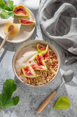 Mısır gevreği veya müsli ile doğal yoğurt, gri arka planda bir kasede incir ve bal, kaşık, meyve ve sabah gölgesi. Kahvaltıda sağlıklı ve besleyici detoks yemeği kavramı. Üst görünüm.