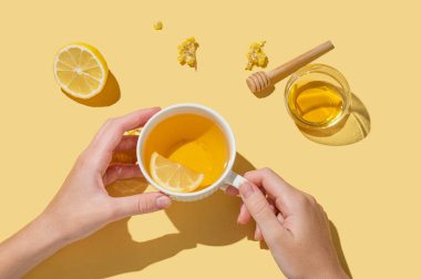 Sarı arka planda, kuru çiçekler ve gölgelerle limon ve bal dolu bir fincan bitki çayını tutan eller. Soğuk mevsimde dokunulmazlık ve sağlığı desteklemek için bir içki konsepti. Üst görünüm, alanı kopyala.