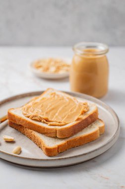 Fıstık ezmeli tost hafif mermer arka planda, fındık ve bıçakla kaplı bir tabakta. Besleyici bir vejetaryen ziyafeti ve Amerikan kahvaltısı için sağlıklı atıştırmalık kavramı. Ön görünüm, boşluğu kopyala.