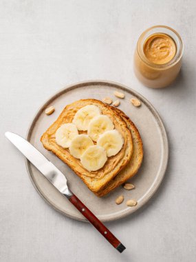 Fıstık ezmeli tost ve muz hafif arka planda, fındık kavanozu, yayılma ve bıçakla birlikte bir tabakta. Amerikan kahvaltısı için besleyici ve sağlıklı atıştırmalıklar. Üst görünüm, alanı kopyala.