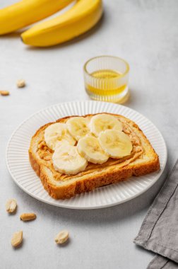 Fıstık ezmeli tost ve muz beyaz bir tabakta bal, meyve ve fındıklı. Amerikan kahvaltısı için besleyici ve sağlıklı atıştırmalıklar. Ön görünüm, boşluğu kopyala.