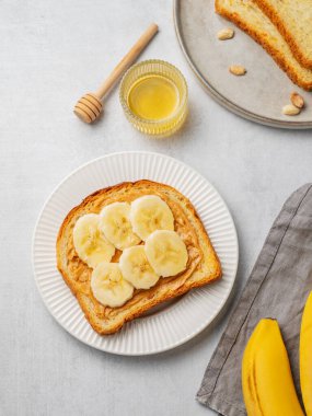 Beyaz bir tabakta bal, meyve ve peçeteyle birlikte fıstık ezmeli tost ve muz. Amerikan kahvaltısı için besleyici ve sağlıklı atıştırmalıklar. Üst görünüm, alanı kopyala.