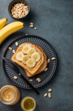 Fıstık ezmeli tost ve muz siyah bir tabakta, fındıklı, serpiştirilmiş ve meyveli. Amerikan kahvaltısı için besleyici ve sağlıklı atıştırmalıklar. Üst görünüm; alanı kopyala.