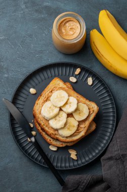 Fıstık ezmeli tost ve muz siyah bir tabakta, fındıklı, serpiştirilmiş ve meyveli. Amerikan kahvaltısı için besleyici ve sağlıklı atıştırmalıklar. Üst görünüm, alanı kopyala.