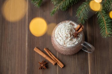 Baharatlar, köknar dalları ve bulanık çelenk ışıklarıyla siyah ahşap arka planda bir fincanda kremalı sıcak çikolata. Sıcak ev yapımı kış içeceği ve tatil atmosferi. Üst görünüm, alanı kopyala.