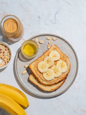 Fıstık ezmeli tost ve muz mermer arka planda meyve, fındık, bal ve sabah gölgesi olan bir tabakta. Amerikan kahvaltısı için tatlı diyet ve sağlıklı atıştırmalıklar kavramı. Üst görünüm, alanı kopyala.