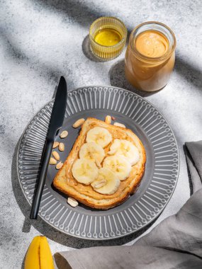 Fıstık ezmeli tost ve muz, gri arka planda, üzerinde bıçak, meyve ve sabah gölgesi olan siyah bir tabakta. Amerikan kahvaltısı için tatlı diyet ve sağlıklı atıştırmalıklar kavramı. Üst görünüm, alanı kopyala.