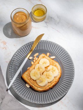 Fıstık ezmeli tost ve muz, bal, bıçak ve sabah gölgesiyle mermer bir zeminde koyu bir tabakta. Amerikan kahvaltısı için tatlı diyet ve sağlıklı atıştırmalıklar kavramı. Üst görünüm, alanı kopyala.