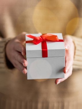 Elinde kırmızı kurdeleli beyaz bir hediye kutusu olan, bej bir süveterin üzerinde yumuşak odaklı ve Noel çelenginden yanıp sönen ışıklar olan bir kadının yakın çekimi. Tatil kavramı ve yeni yıl konforu. Ön görünüm.