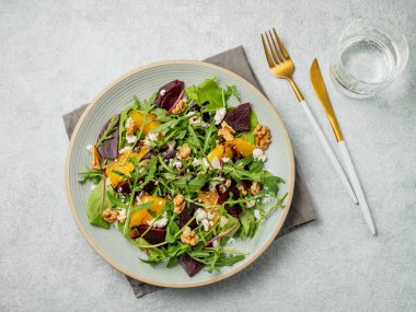 Kavrulmuş pancar, portakal ve roka salatası, beyaz peynir, ceviz ve hafif bir arka planda hafif bir tabakta balzamik sos, çatal bıçak takımı ve bir bardak su. Sağlıklı organik gıda konsepti. Üst görünüm, alanı kopyala.