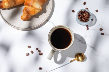 Kruvasan, kahve çekirdeği, kaşık ve güneş ışığıyla beyaz mermer bir arka planda taze demlenmiş kahve. Lezzetli kahvaltı kavramı. Üst görünüm, alanı kopyala.