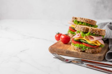 Jambonlu, peynirli, marullu, sebzeli ve tahtadan ekmek serpiştirilmiş iki sandviç mermer arka planda, çatal bıçaklı ve peçeteli. Lezzetli, hızlı ve sağlıklı bir kahvaltı. Ön görünüm, boşluğu kopyala.