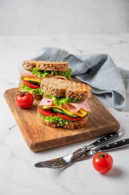 Jambonlu, peynirli, marullu, sebzeli ve tahtadan ekmek serpiştirilmiş iki sandviç mermer arka planda, çatal bıçaklı ve peçeteli. Lezzetli, hızlı ve sağlıklı bir kahvaltı. Ön görünüm, boşluğu kopyala.