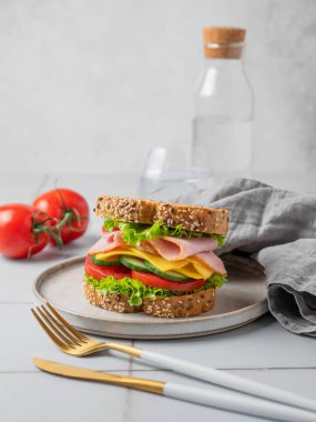 Sandviç ya da kızarmış ekmek, jambon, peynir, marul ve sebzeler gri fayanslı bir tabakta ve hafif arka planda çatal bıçak, bardak su ve peçeteyle. Sağlıklı ev atıştırmalığı konsepti. Ön görünüm, boşluğu kopyala.