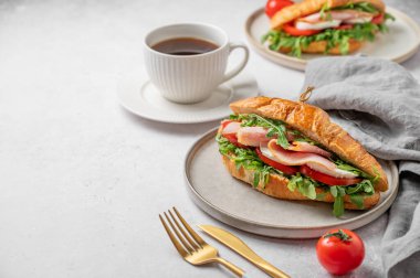 Jambonlu kruvasan sandviçi, mozzarella peyniri, domates, bir tabak roka ve arka planda hafif arkaplanda bir fincan kahve, içinde de Cutlry ve peçete. Sağlıklı bir kahvaltı ya da brunch kafe menüsü. Ön görünüm, boşluğu kopyala.