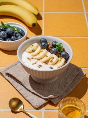 Turuncu bir zemin üzerinde bal, meyve, böğürtlen ve sabah güneş ışığıyla turuncu bir kapta yaban mersini ve muzlu doğal Yunan yoğurdu. Sağlıklı ve besleyici kahvaltı konsepti. Ön görünüm, boşluğu kopyala.