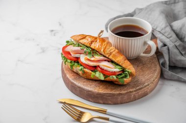 Jambonlu kruvasan sandviç, mozzarella peyniri, domates, ahşap bir tahtanın üzerinde roka ve mermer arka planda çatal bıçaklı bir fincan kahve. Sağlıklı bir kahvaltı ya da brunch kafe menüsü. Ön görünüm, boşluğu kopyala.