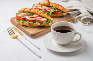 Jambonlu kruvasan sandviçi, mozzarella peyniri, domates, ahşap bir tahtada roka ve mermer arka planda çatal bıçaklı bir fincan kahve. Sağlıklı bir kahvaltı ya da brunch kafe menüsü. Ön görünüm.