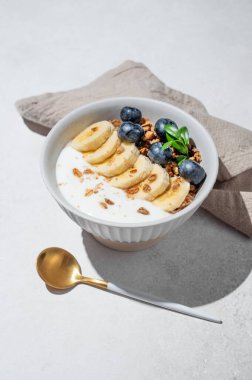Doğal Yunan yoğurdu, granola, yaban mersini ve muz ile bez, kaşık ve sabah gölgeleriyle birlikte beton bir arka planda. Sağlıklı ve besleyici kahvaltı konsepti. Ön görünüm, boşluğu kopyala.