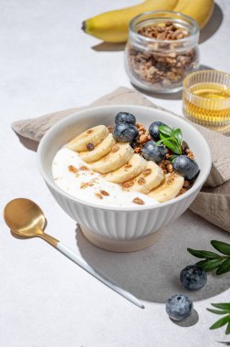 Bal, meyve ve sabah gölgeleriyle birlikte açık beton arka planda bir kasede yaban mersini ve muzlu doğal Yunan yoğurdu. Sağlıklı ve besleyici kahvaltı konsepti. Ön görünüm, boşluğu kopyala.