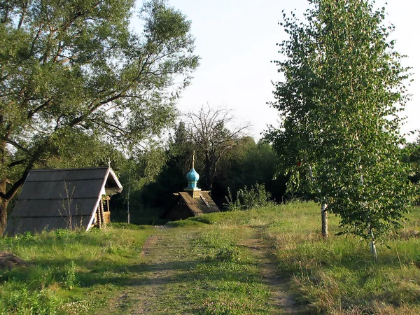 Un camino de campo conduce a una estructura de madera y una capilla ortodoxa, que se encuentran entre los árboles. — Foto de Stock