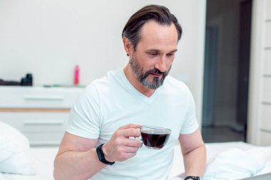 Kendine güvenen, sakallı, beyaz bir adam evde oturmuş bir fincan kahveyle gülümsüyor.