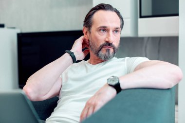 Sakallı, sakallı, sakallı, sakin bir adam, kanepede oturup uzaktan bakarken boynuna dokunuyor.