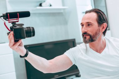 Yakışıklı, tıraşsız, genç bir adam kamerayla elini öne koyuyor ve kendinden emin görünüyor.