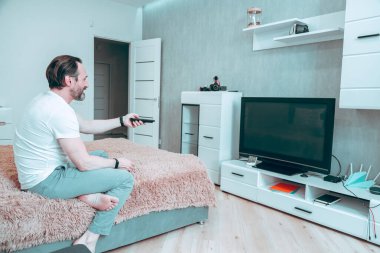 Tatmin olmuş çıplak ayaklı adam uzaktan kumandayla TV ekranına bakıp gülümsüyor.