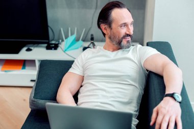 Beyaz tişörtlü neşeli, çekici beyaz adam dizüstü bilgisayarla yatarken uzaktan bakıp gülümsüyor.